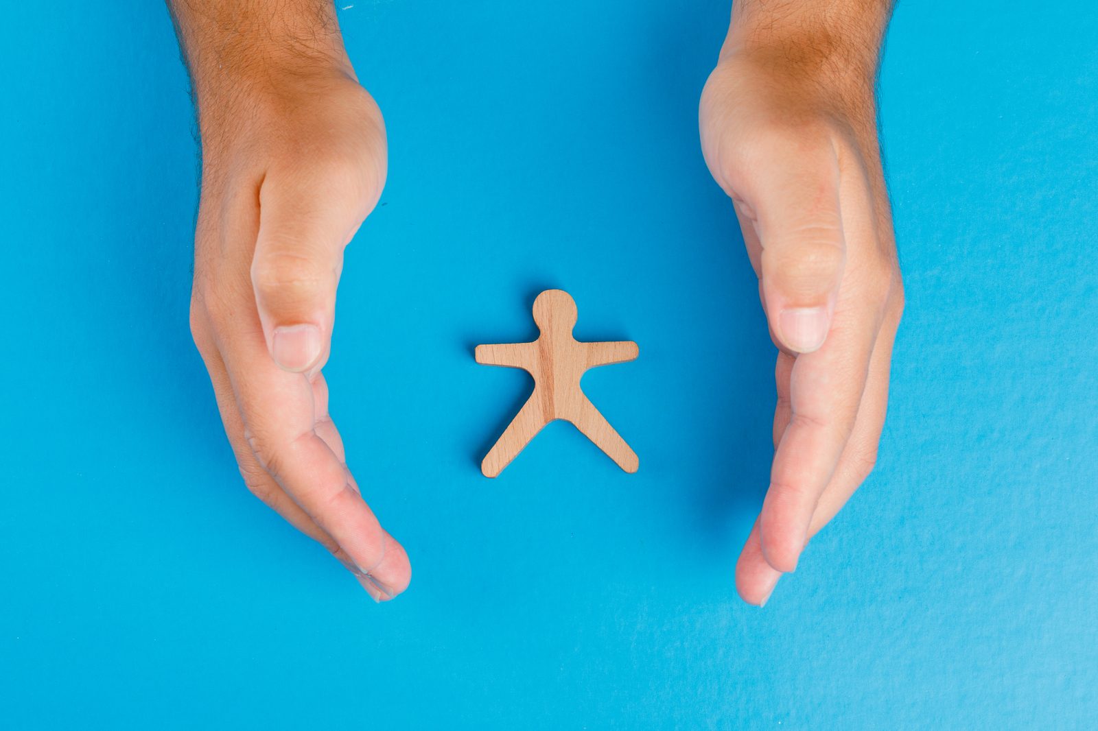 Two hands cradling a small wooden human figure on a blue background, suggesting protection and oversight